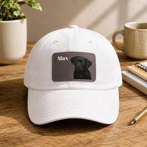 May include: A white baseball cap featuring a rectangular patch on the front. The patch is dark gray with a stitched border and displays a portrait of a black dog with the name Max printed in white text to the left of the dog's head.