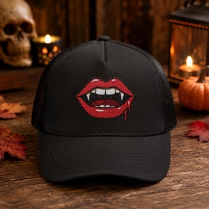 May include: A black trucker cap featuring an embroidered graphic of red lips with white vampire fangs and a single drop of red blood dripping from the corner of the mouth. The cap has a solid front panel and a mesh back panel construction.