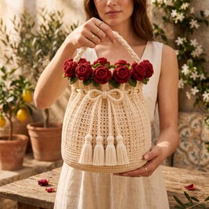May include: A beige crochet bucket bag featuring a braided handle and a drawstring closure with four long tassels. The top edge is decorated with a row of deep red crocheted roses with green leaves, creating a floral border around the opening.