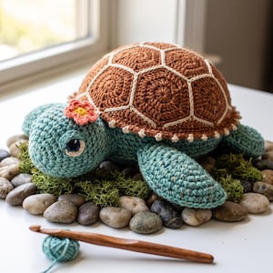 Può includere: Un peluche a forma di tartaruga marina realizzato all'uncinetto con un corpo testurizzato color verde acqua e un guscio marrone con motivi esagonali cuciti in bianco. Un piccolo fiore all'uncinetto rosa e giallo è fissato sulla testa.