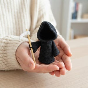 May include: A small, black crocheted figure wearing a hooded cloak, holding a miniature scythe with a light-colored wooden handle and a white blade. The figure is crafted with a textured yarn stitch and features a deep, dark hood.