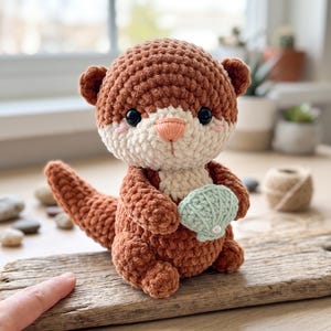 May include: A brown and cream-colored crochet otter plush toy with black eyes, a peach-colored nose, and pink blush marks. The otter is holding a small, light green crocheted seashell with a single white pearl bead attached to the center.