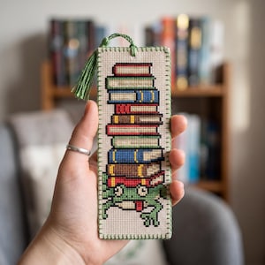 May include: A rectangular cross-stitch bookmark on light beige fabric featuring a stack of colourful books balanced on the head of a green frog. The bookmark is edged with green stitching and includes a green tassel at the top, showcasing detailed embroidery.