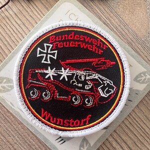 Könnte beinhalten: Ein kreisförmiger gestickter Aufnäher mit schwarzem Hintergrund und einem rot-gelb-schwarzen Rand. Er zeigt eine Grafik eines rot-weißen Feuerwehrautos, ein weißes Kreuz sowie den roten Text "Bundeswehr Feuerwehr" und "Wunstorf" in Fettschrift.