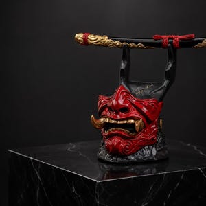 May include: A red and black oni mask sculpture featuring gold-colored fangs and a textured base. The mask serves as a stand for a black sword scabbard, which is decorated with gold-colored metal accents and tied with a red cord at the top.