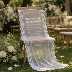 May include: A sheer white fabric memorial sign draped over a wooden chair, featuring white lace trim and the text: RESERVED IN MEMORY, This place is lovingly reserved for someone who cannot be with us today, Margaret Wilson, BELOVED GRANDMA OF THE GROOM.