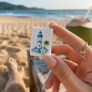 May include: A rectangular white tile featuring a colorful illustration of a blue and white lighthouse, a green palm tree, and a white tropical flower. The tile is held against a backdrop of a sandy beach, ocean waves, and a coconut drink.