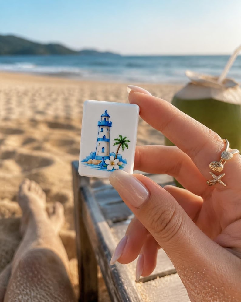 May include: A rectangular white tile featuring a colorful illustration of a blue and white lighthouse, a green palm tree, and a white tropical flower. The tile is held against a backdrop of a sandy beach, ocean waves, and a coconut drink.