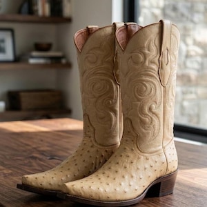 May include: A pair of tan cowboy boots featuring a textured ostrich leather vamp with distinct quill patterns and a smooth leather shaft adorned with intricate tonal stitching. The boots have a pointed toe, a stacked heel, and pull tabs.