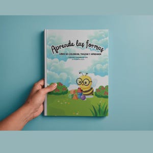 Puede incluir: Un libro de actividades de tapa blanda titulado Aprende las formas, Libro de colorear, trazar y aprender. La portada muestra una abeja de dibujos animados con gafas, césped verde, arbustos y formas geométricas coloridas bajo un cielo azul.