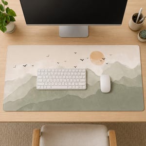 Peut inclure: Un tapis de bureau rectangulaire présentant un paysage à l'aquarelle composé de montagnes superposées vert sauge, d'un ciel beige pâle, d'un soleil orange doux et de petites silhouettes d'oiseaux noirs en vol sous un clavier et une souris.