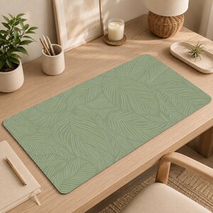 May include: A rectangular desk mat featuring a sage green background with a repeating pattern of white line-drawn tropical leaves. The mat has a smooth surface and rounded corners, providing a flat area for workspace organization and protection.