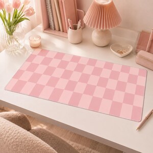 Puede incluir: Una alfombrilla de escritorio rectangular con un patrón de cuadros repetitivo en dos tonos de rosa claro. La alfombrilla está colocada sobre un escritorio blanco, rodeada por una lámpara rosa, un jarrón con tulipanes rosas, una vela, un portalápices y un pequeño plato para joyas.