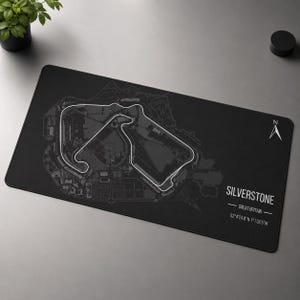 Op de afbeelding: Een rechthoekige zwarte bureaumat met een witte omtrek van het Silverstone-racecircuit in Groot-Brittannië. Het ontwerp bevat een noordpijl-icoon, de tekst SILVERSTONE, GREAT BRITAIN en de coördinaten 52°4'24.8"N 1°1'32.5"W.