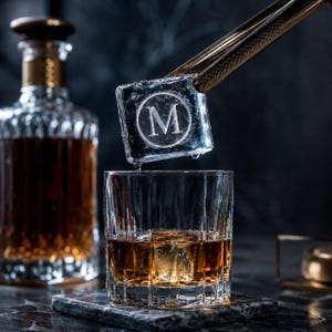 Peut inclure: Un glaçon carré transparent gravé de la lettre M dans un cercle, tenu par une pince en métal doré au-dessus d'un verre en cristal rempli d'un liquide ambré. Une carafe en cristal assortie remplie de liquide ambré se trouve en arrière-plan.