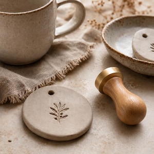 May include: A round, light beige clay ornament featuring an embossed olive branch design and a small hole at the top. Beside it sits a brass wax seal stamp with a smooth, light-colored wooden handle, along with a speckled ceramic mug and bowl.