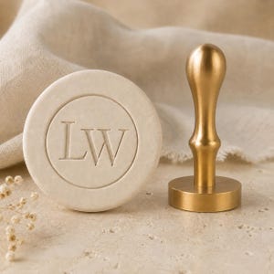May include: A circular off-white wax seal with the embossed letters LW in the center, positioned next to a gold-toned metal stamp with a smooth, rounded handle and a flat circular base used for creating wax impressions.
