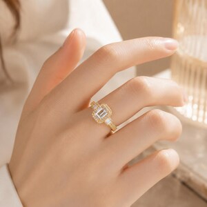 Könnte beinhalten: Ein goldener Ring mit einem zentralen, smaragdgeschliffenen, klaren Stein, der von einem Kranz aus kleinen runden Steinen umgeben ist, sowie zwei zusätzlichen Baguette-geschliffenen Steinen an den Seiten der Ringschiene aus poliertem Gold.