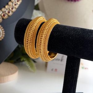 Può includere: Due bracciali rigidi color oro caratterizzati da una superficie testurizzata molto dettagliata, con bordi a perline complessi e un motivo centrale ripetitivo. I bracciali hanno un profilo spesso e arrotondato con finitura metallica lucida.