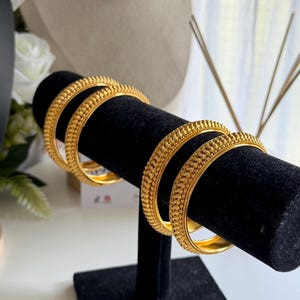 May include: A pair of gold-toned bangles featuring a textured, beaded surface design. The circular bracelets have a polished interior and a detailed, raised pattern along the outer edge, displayed on a black cylindrical stand.