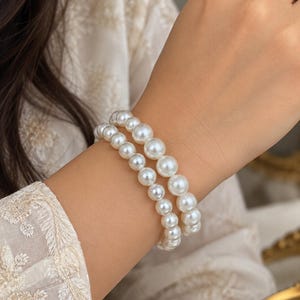 May include: A double-strand bracelet featuring white, spherical faux pearls of varying sizes. The larger pearls are positioned in the centre of the strands, gradually tapering to smaller pearls towards the ends, creating a graduated design.