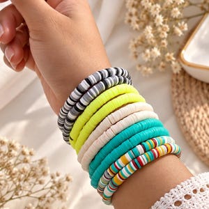 May include: A stack of five bracelets made from flat, circular polymer clay beads. The bracelets feature various color patterns, including a black, gray, and white mix, solid neon yellow, solid cream, solid turquoise, and a multicolored striped design.