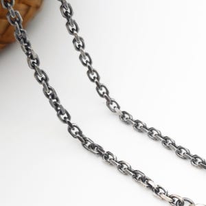 Pode incluir: Um colar de corrente de metal em tom prateado escuro com elos ovais e um acabamento oxidado texturizado. A corrente inclui um fecho lagosta numa extremidade e um pequeno anel de ligação circular na outra, com um padrão de elos consistente.