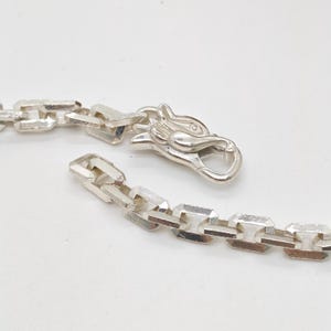May include: A silver-toned chain necklace featuring rectangular, faceted links with a textured finish. The necklace includes a decorative clasp shaped like a stylized animal head, which serves as the functional closure mechanism for the chain.