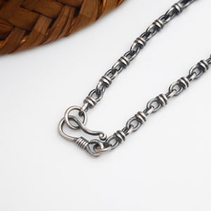 Op de afbeelding: Een zilverkleurige metalen ketting bestaande uit een reeks onderling verbonden ovale schakels met decoratieve geribbelde banden. De ketting wordt vastgemaakt met een gebogen haaksluiting die aan een ronde ring bevestigd is.