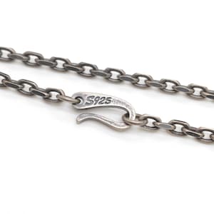 May include: A dark, oxidised silver chain necklace featuring oval-shaped links and a hook-style clasp. The metal clasp is clearly stamped with the text S925, indicating the material composition. The chain has a weathered, matte finish throughout.