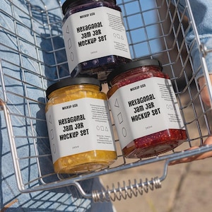 May include: Three hexagonal glass jars with black lids, each containing 240ml of fruit preserves in yellow, red, and dark purple. Each jar features a white label with the text Hexagonal Jam Jar Mockup Set and Mockup Asia, held in a wire basket.