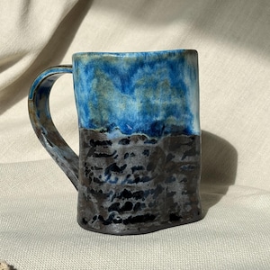 Pode incluir: Uma caneca de cerâmica com um design de esmalte de dois tons. A parte superior é de um azul vibrante e brilhante com tons verdes subtis, enquanto a parte inferior é de um cinzento-carvão escuro e texturizado com um aspeto manchado e terroso.