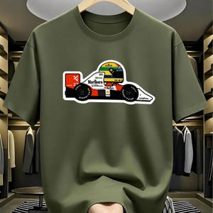May include: An olive green short-sleeve t-shirt featuring a graphic of a Formula One race car with a driver wearing a yellow, green, and blue helmet. The car displays the Marlboro, Honda, and Shell logos, the number 27, and Goodyear tire branding.