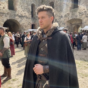 May include: A dark charcoal gray hooded cloak featuring a faux fur collar and brown leather buckle straps. The garment is worn over a dark leather chest piece with additional leather straps and a sword hilt visible at the waist area.
