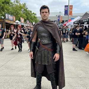 May include: A dark brown hooded cloak worn over a dark grey chest plate and a matching skirt with vertical panels. The outfit includes dark brown trousers, black arm bracers, black boots, and a sword with a brown belt worn across the torso.