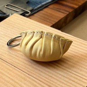 Puede incluir: Un pequeño llavero de cuero amarillo con forma de empanadilla, con un anillo plateado. El llavero está sobre una superficie de madera de color claro. La empanadilla tiene un diseño plisado y está cosida a lo largo del borde superior.