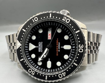 May include: A Seiko automatic wristwatch with a black dial, luminous circular hour markers, and a rotating black bezel with white numerals. The watch features a day-date window at the three o'clock position, a stainless steel jubilee bracelet, and text reading Seiko Automatic, Diver's 200m, 21 Jewels, and Made in Japan.