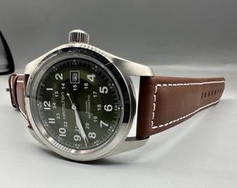Op de afbeelding: Een Hamilton Khaki Automatic horloge met een ronde roestvrijstalen kast, een olijfgroene wijzerplaat met witte Arabische cijfers, een datumvenster op de drie-uurpositie en een bruine leren band met witte contraststiksels.