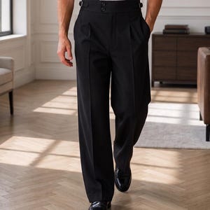 Puede incluir: Pantalones negros de talle alto con silueta de pierna ancha, cintura estructurada con dos botones frontales y trabillas de ajuste laterales. Los pantalones incluyen pliegues frontales profundos y un diseño recto con un pliegue vertical marcado.