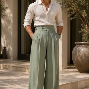 May include: A beige long-sleeved button-down shirt paired with high-waisted, wide-leg sage green trousers featuring a double-button closure, side adjusters, and a pleated front. The outfit is completed with light-colored loafers and dark sunglasses.