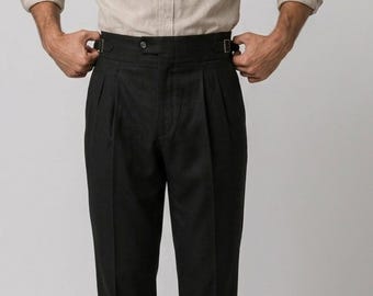 May include: Black high-waisted trousers featuring double pleats at the front, a wide waistband with side adjusters, and a button closure. The trousers are paired with a light beige long-sleeved button-down shirt tucked into the waistband.