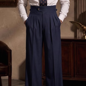 May include: High-waisted navy blue trousers featuring a wide-leg silhouette, double pleats at the front, and a structured waistband with side adjusters. The trousers are paired with a white long-sleeved button-down shirt and a dark patterned tie.