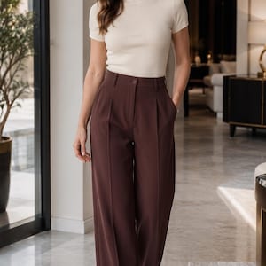 Puede incluir: Una blusa de manga corta en tejido de punto acanalado color crema con cuello alto, combinada con pantalones de talle alto en color marrón oscuro de pierna ancha, con cierre de botón, trabillas para cinturón y pliegues verticales.