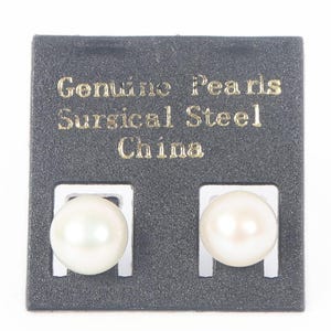May include: A pair of white round pearl stud earrings mounted on a black display card. The card features gold text that reads Genuine Pearls, Sursical Steel, and China. The earrings are positioned side by side on the textured black backing.
