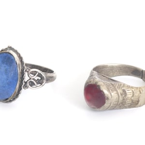 May include: Two metal rings. The left ring features a large, oval, deep blue stone set in a twisted metal frame with a decorative openwork band. The right ring has a smooth, rounded, translucent red stone set in a band with etched line details.