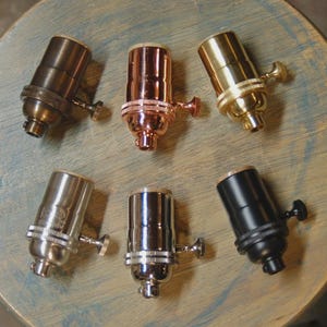 May include: Six cylindrical metal light bulb sockets arranged in two rows. The finishes include antique bronze, polished copper, brushed brass, brushed nickel, polished chrome, and matte black. Each socket features a side-mounted turn knob switch.