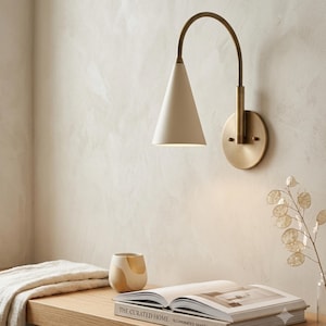 May include: A wall-mounted lamp featuring a curved brass arm and a matte cream-coloured conical shade. The fixture includes a circular brass wall plate. The lamp is positioned above a wooden surface holding a book, a small ceramic cup, and a plant.
