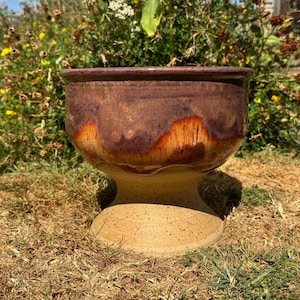 May include: A ceramic bowl with a wide, flared rim and a pedestal base. The upper portion features a glossy, deep plum and burnt orange glaze with a drip effect, while the lower pedestal base is a matte, speckled beige clay finish.