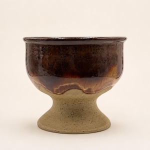 May include: A ceramic chalice with a glossy, dark brown glazed upper bowl and a matte, speckled beige base. The vessel features a wide, rounded rim and a sturdy, flared pedestal foot, showcasing a distinct contrast between the smooth dark glaze and the textured, light-colored clay body.