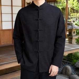 Pode incluir: Uma camisa preta de manga comprida com gola mandarim e fecho frontal fixado por cinco botões de alternância revestidos de tecido. A peça de vestuário tem uma bainha reta e uma cor sólida e uniforme em toda a sua extensão.
